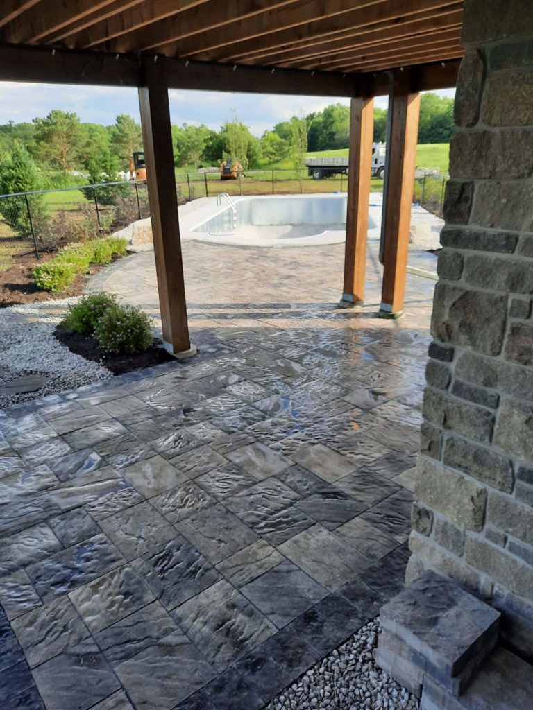 View from a shaded stone patio with textured paving, looking out towards a swimming pool surrounded by greenery and another small shelter.