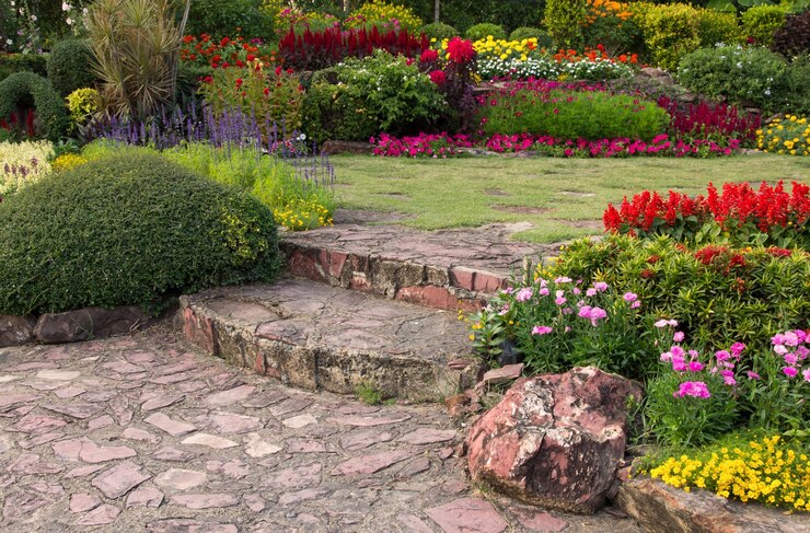 A well-manicured garden with a stone path leading through vibrant flower beds of red, purple, yellow, and pink blooms, complemented by lush green shrubs.